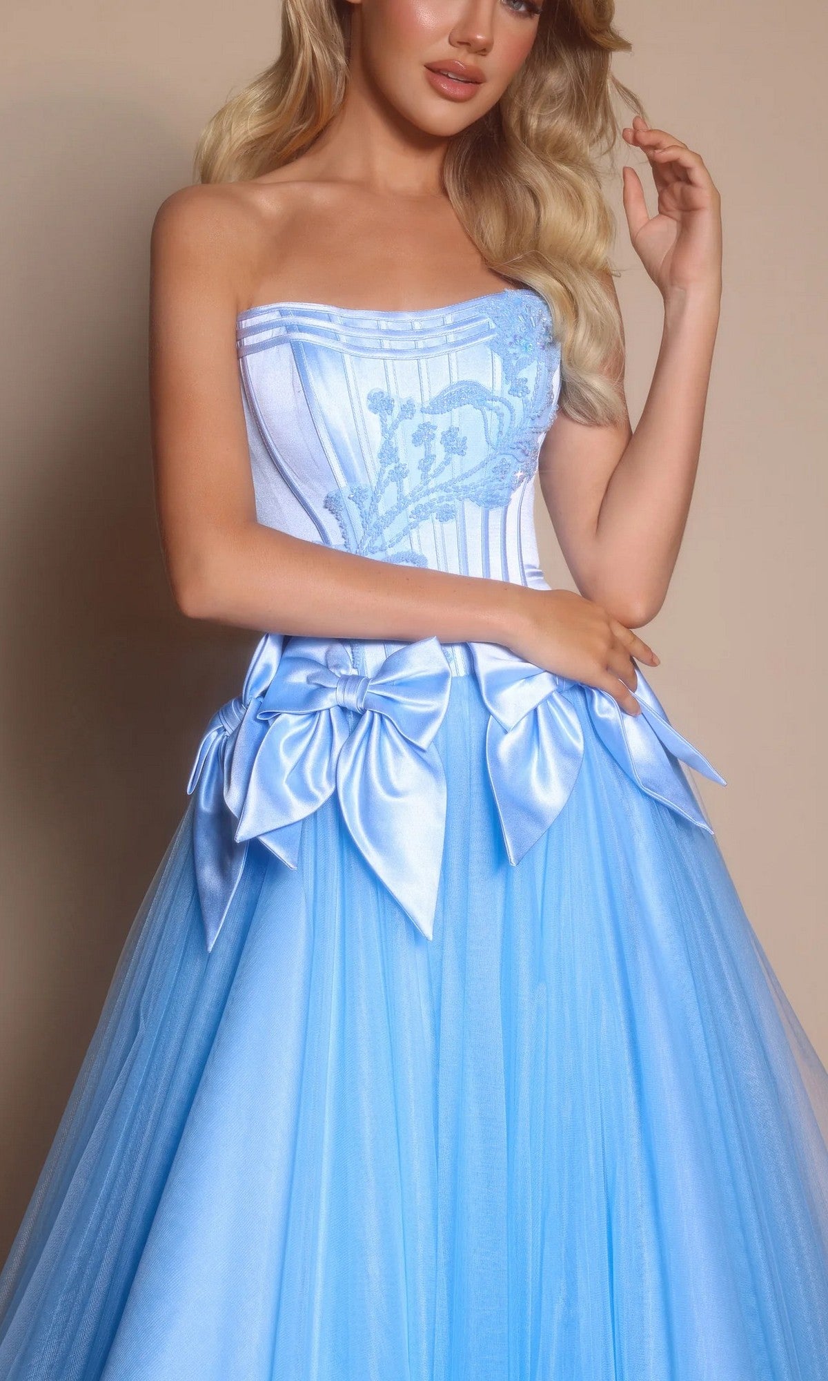 A woman wears the Strapless Ball Gown: Portia and Scarlett PS26102, featuring a light blue fitted corset bodice, blue embroidery, and large satin bows at the waist.
